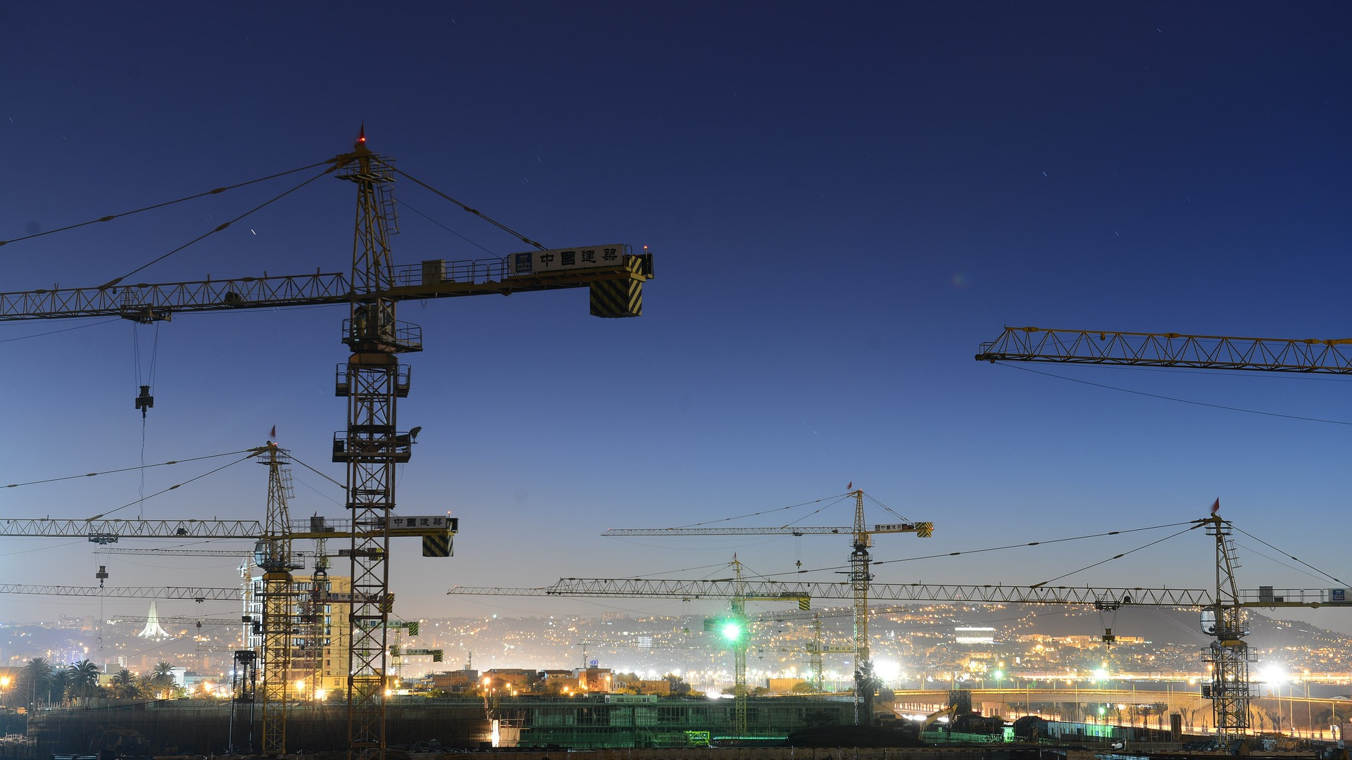 A cluster of towering construction cranes set against a sky at dusk with distant city lights in the background